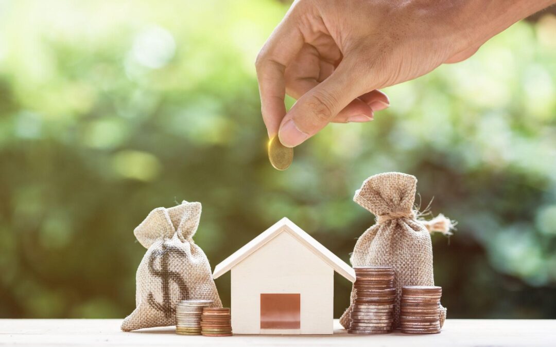 A person holds a coin over a tiny model of a home, which is surrounded by small bags of coins.